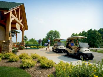 People in golf carts are near a building with wooden beams and greenery in the foreground. Another person is walking with a golf bag.