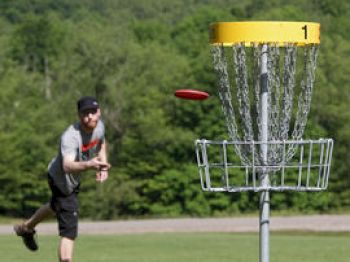 A person is playing disc golf, aiming a frisbee towards a metal basket with chains, set in a grassy field surrounded by trees.