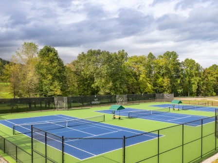 This image shows an outdoor tennis court area with blue and green courts, surrounded by a black fence and trees. Two green umbrellas are present.