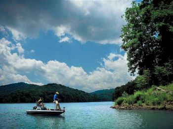 Two people are fishing on a boat in a serene lake surrounded by forested hills under a partly cloudy sky.