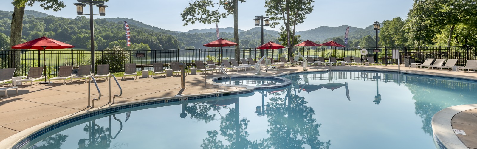 An outdoor swimming pool with clear water, surrounded by lounge chairs and red umbrellas. In the background are trees and mountains.
