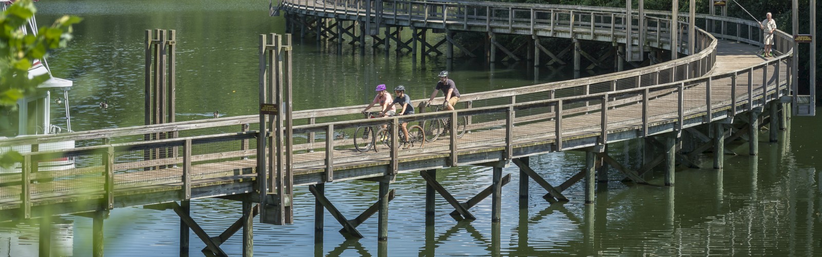 A serene lakeside scene with a winding wooden boardwalk over the water, surrounded by lush greenery, and a few people walking.