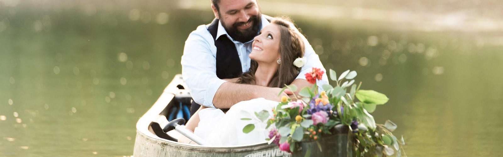 A couple is sitting in a canoe on a calm lake, smiling and embracing each other, with a bouquet of flowers in the canoe.