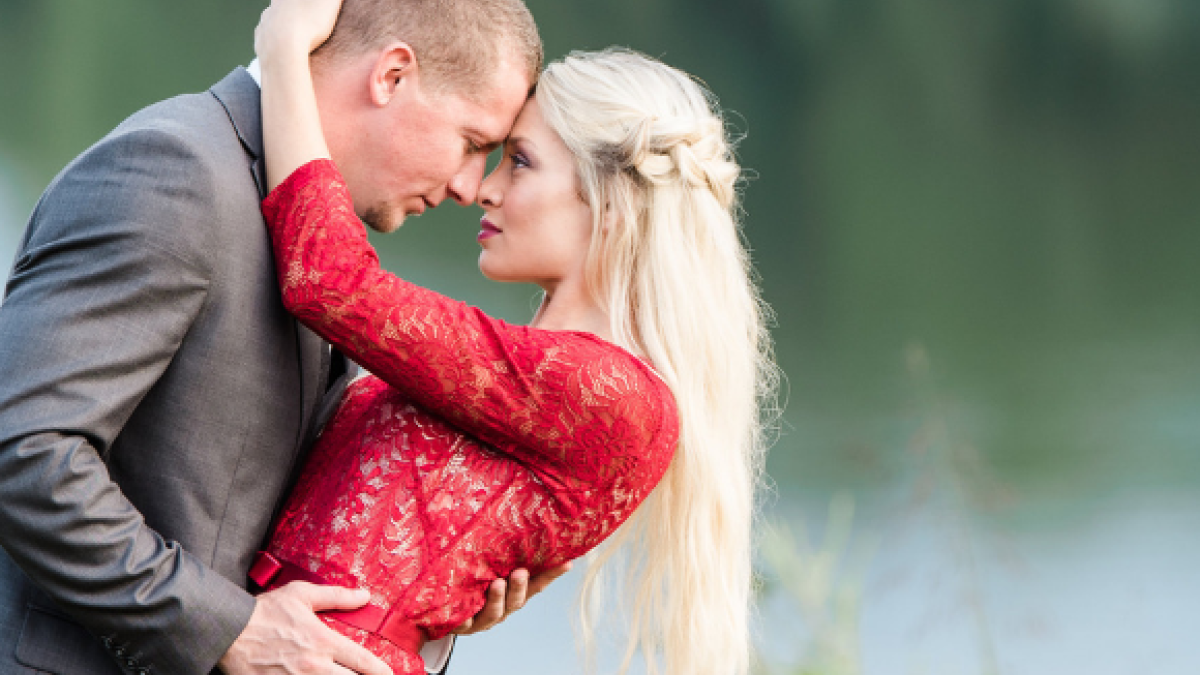 A couple is embracing near a body of water; the man in a suit and the woman in a red dress, with greenery in the background.