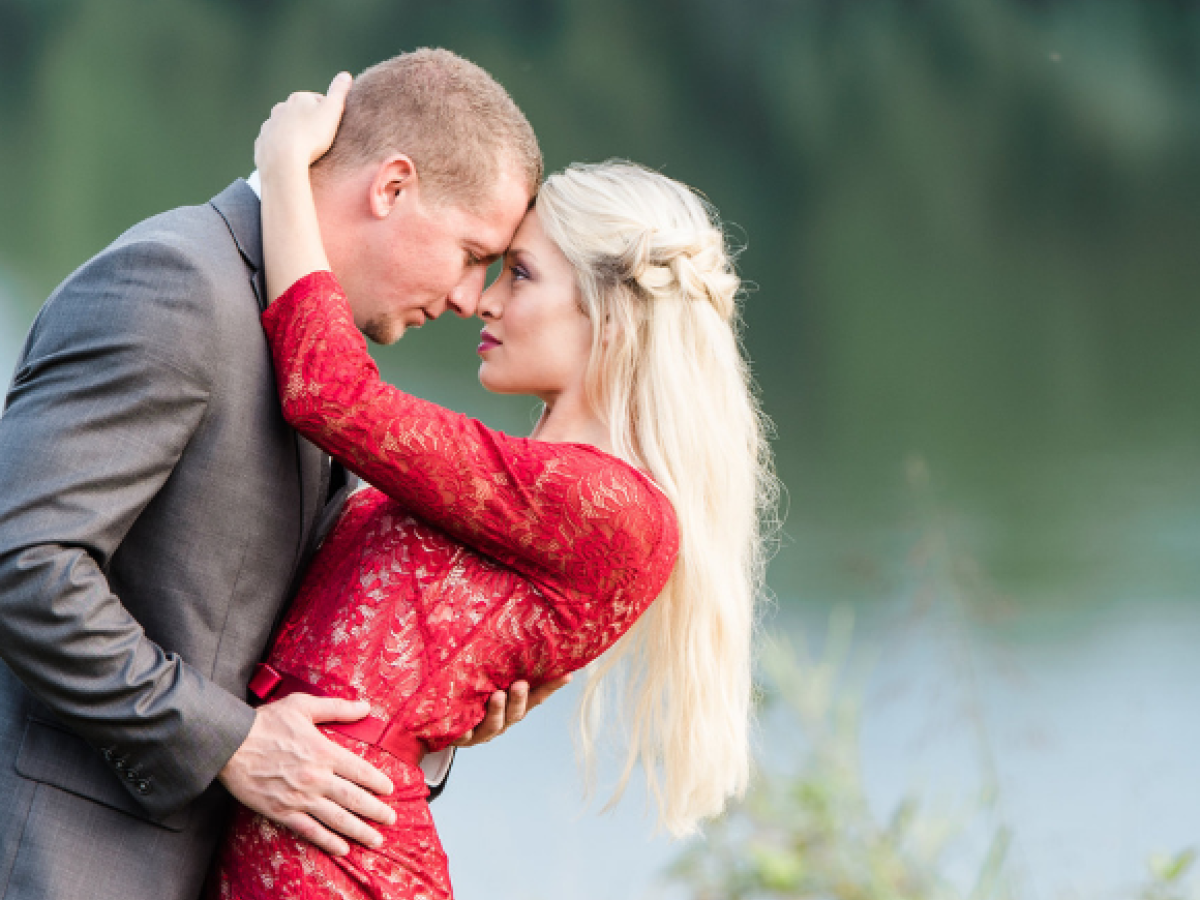A couple is embracing near a body of water; the man in a suit and the woman in a red dress, with greenery in the background.