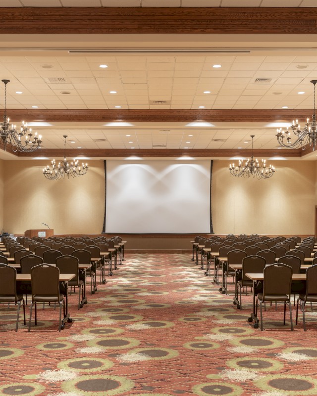 An empty conference or presentation room with rows of chairs facing a large projection screen at the front.