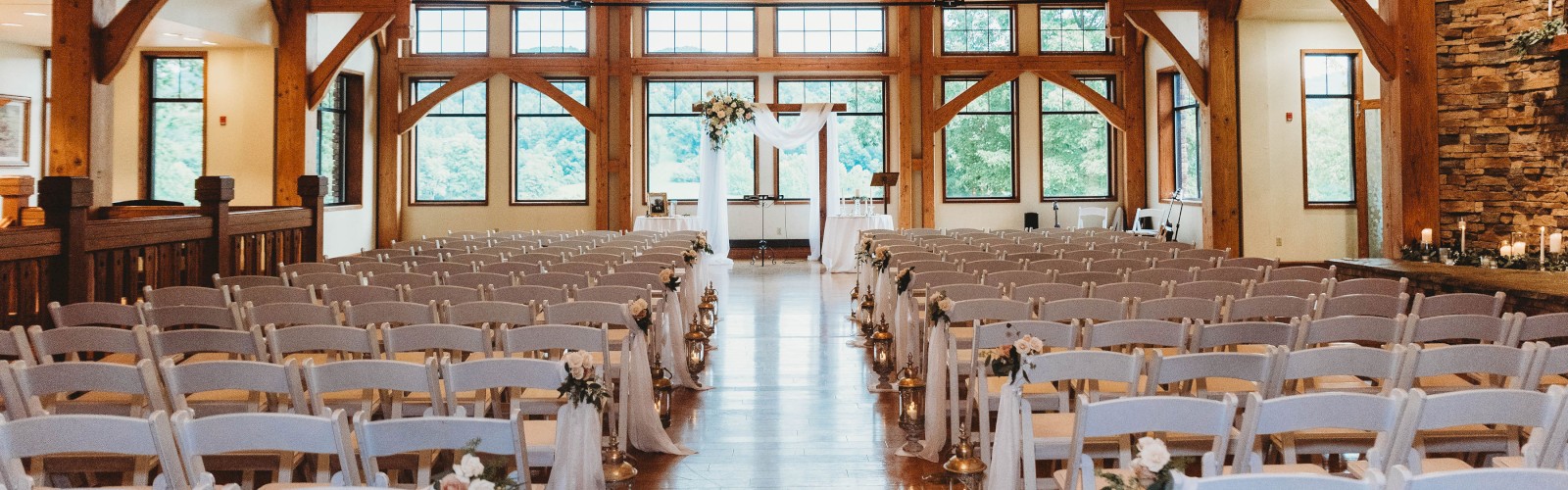 This image shows a beautifully decorated wooden wedding venue with rows of white chairs arranged facing an altar, and chandeliers overhead.