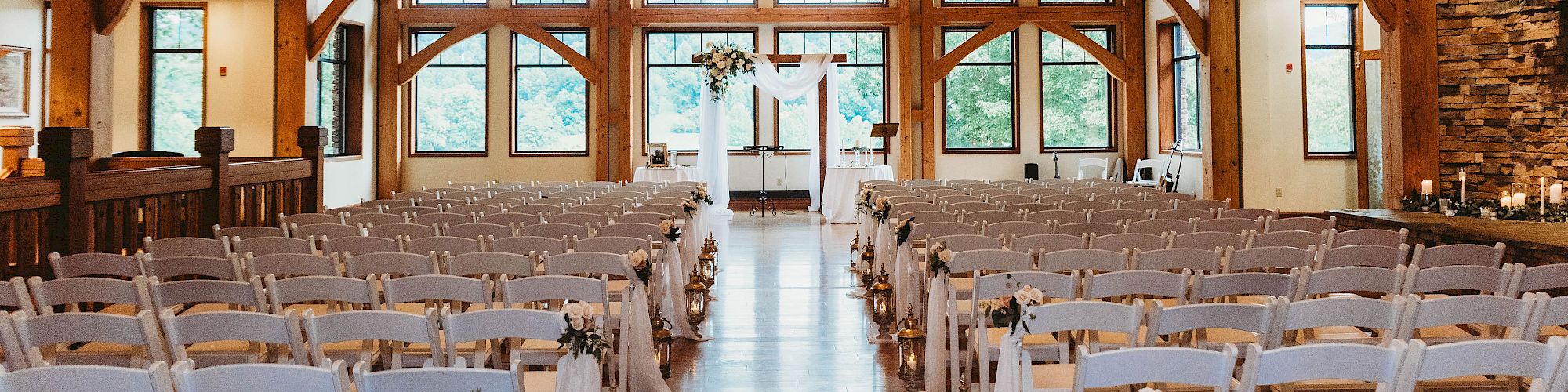 This image shows a beautifully decorated wooden wedding venue with rows of white chairs arranged facing an altar, and chandeliers overhead.