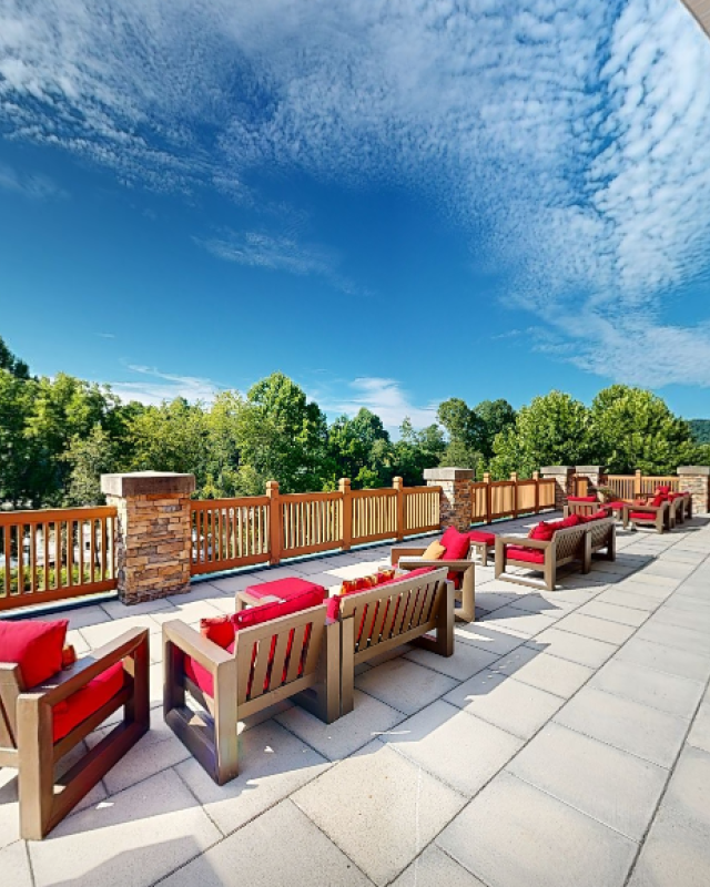 A spacious outdoor terrace with wooden chairs and red cushions overlooks lush green trees and a blue sky with wispy clouds.