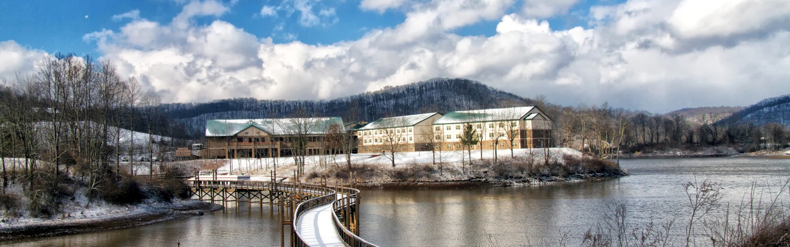 A serene landscape with buildings by the water, a curved snowy walkway leading to them, surrounded by mountains and a partly cloudy sky.