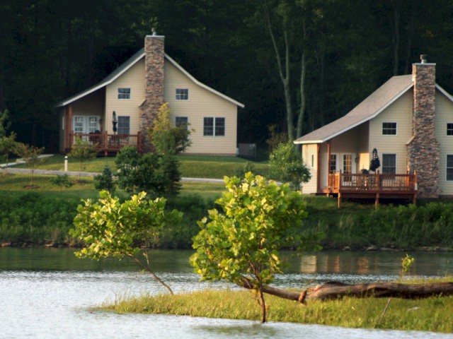 Two cream-colored houses with stone chimneys are next to a river, surrounded by green trees and a grassy area with small trees.