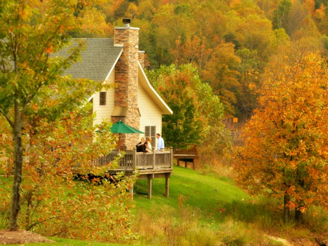 A house with a deck, surrounded by colorful autumn trees and forested hills in the background.