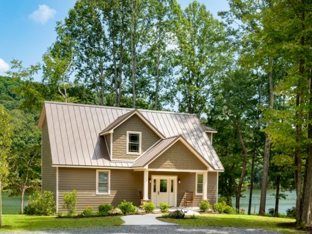 A modern house with a metal roof, surrounded by trees and a well-maintained lawn, with a lake visible in the background on a sunny day.