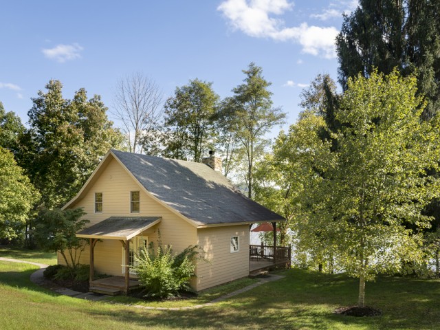 A cozy beige house surrounded by green trees and a lush lawn under a partly cloudy sky.