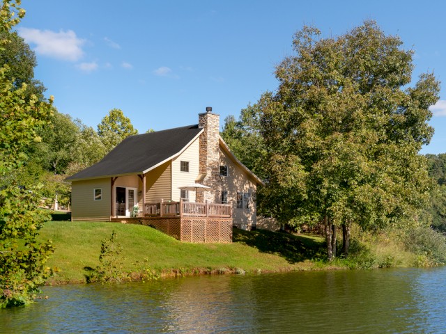 A cozy house by a lake with lush trees, a green lawn, clear blue sky, and a shaded deck area.