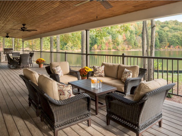 A large outdoor patio with wicker furniture, flowers, and a view of a lake surrounded by trees in fall.