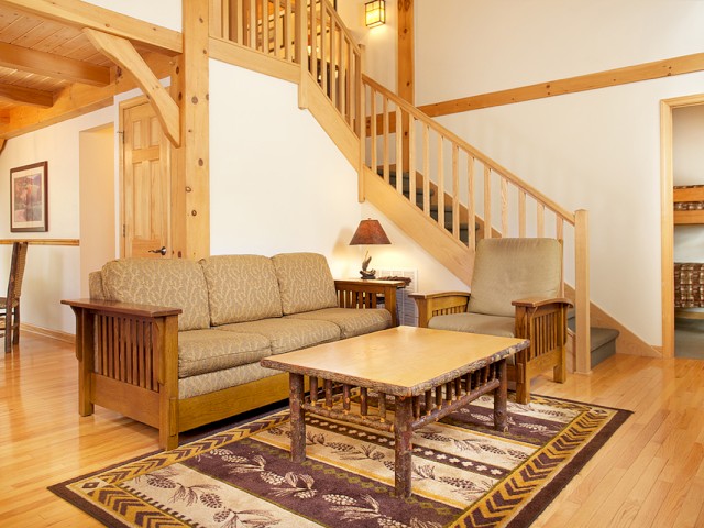 A cozy living room with wooden furniture, a staircase, and a rug, leading to a room with bunk beds, all in a warm, rustic style.