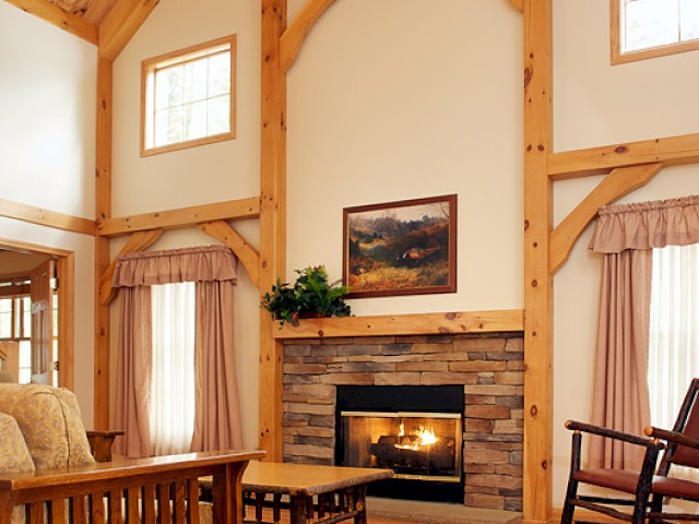 A cozy living room with a stone fireplace, wooden beams, and furniture, illuminated by natural light from small windows.