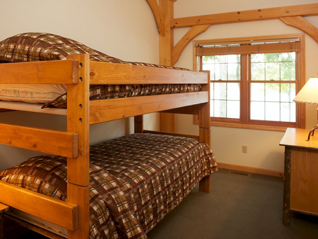 A cozy room with a wooden bunk bed, a window with blinds, and a small lamp on a wooden dresser.