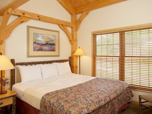 A cozy bedroom with a rustic wooden bed frame, matching bedside tables, a pair of lamps, a colorful floral quilt, and large windows with wooden blinds.