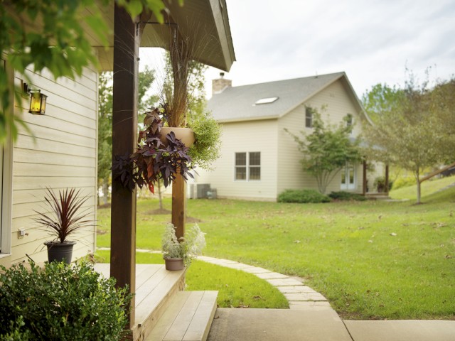 This image shows a cozy backyard with a porch, hanging plants, potted plants, and a curved stone pathway leading to a house.