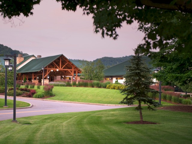 A scenic view of a well-maintained grassy area with trees, a winding road, a large pavilion, and mountain background at sunset.