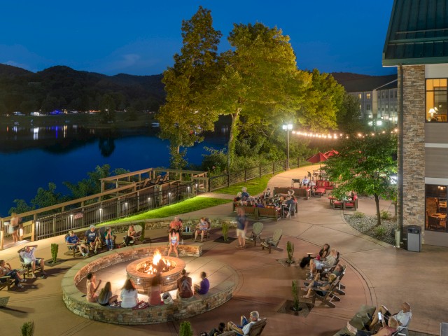 People gather around a fire pit on a patio near a lake, with trees, buildings, and mountains in the background at dusk.