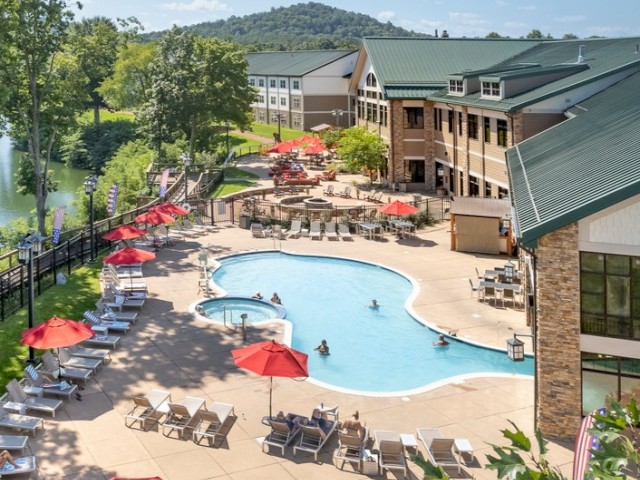 A resort with a large outdoor pool, red umbrellas, lounge chairs, a hot tub, and a nearby building with a green roof, surrounded by trees and hills.
