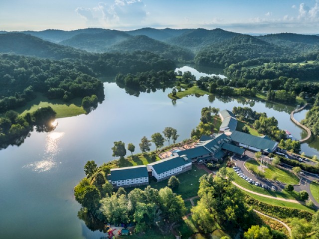 This image shows a large, modern building complex next to a winding lake, surrounded by lush green trees and rolling hills on a sunny day.
