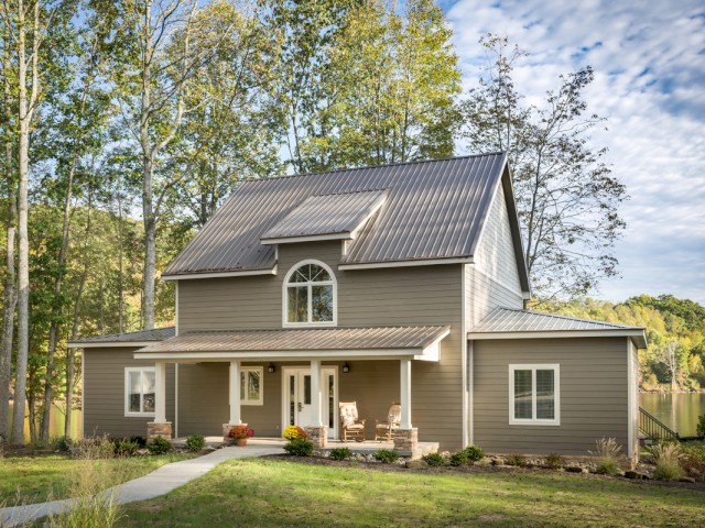 A modern house with a steep metal roof, porch, and large windows, built on a lush green lawn with trees and a water body nearby.