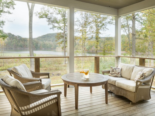 A cozy porch with wicker furniture, cushions, and a table with drinks overlooks a scenic lake and wooded landscape.