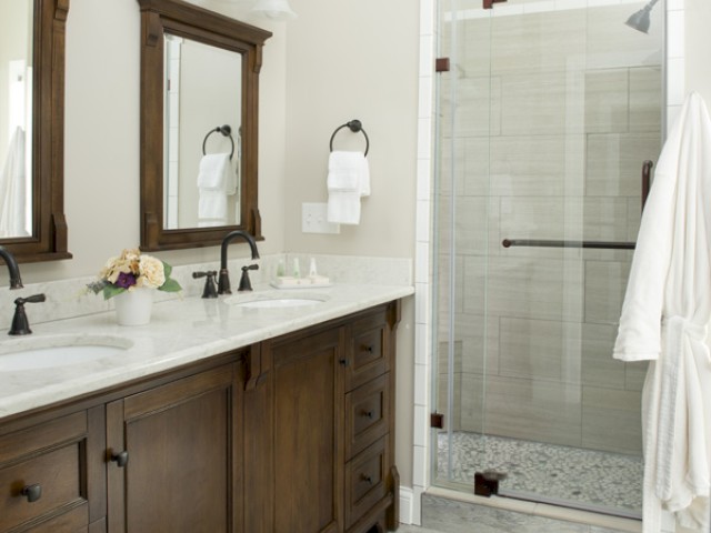 The image shows a bathroom with a double vanity, wooden cabinets, a marble countertop, and a glass-enclosed shower with a towel hanging.