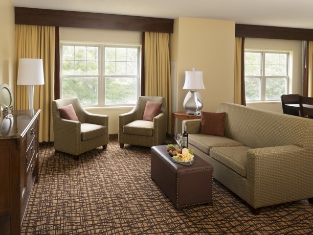 A cozy hotel living area with chairs, a sofa, lamps, a TV, and a window view of trees.