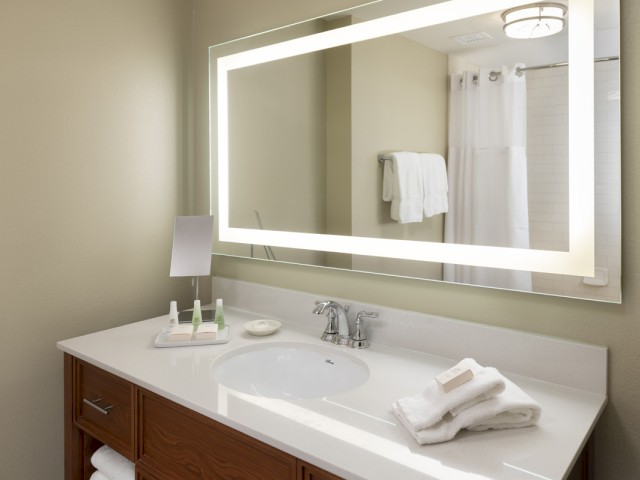 A clean bathroom vanity with towels, toiletries, and a large illuminated mirror reflecting the shower area.