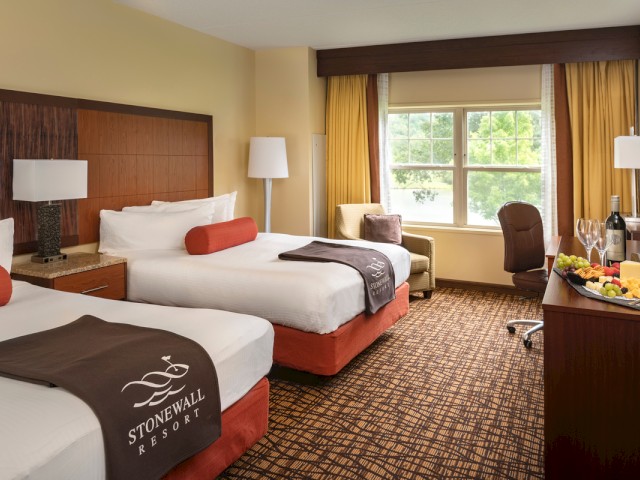 A hotel room with two beds, a window view of greenery, a desk with snacks and drinks, and cozy furniture at Stonewall Resort.