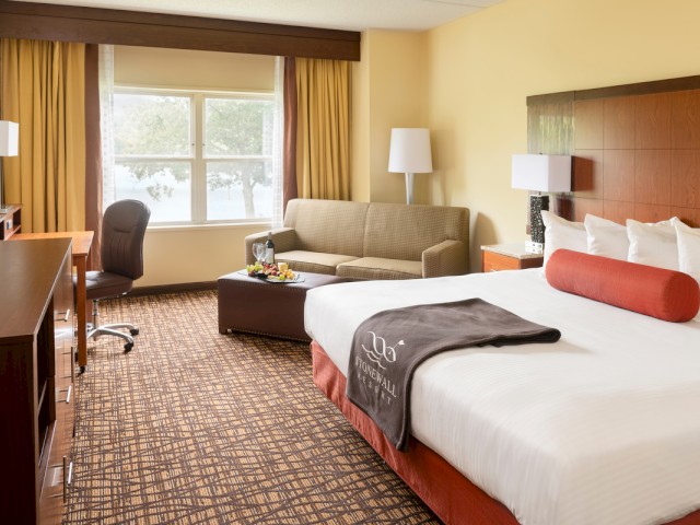 This image shows a hotel room with a large bed, a sofa, a desk, a TV, and a window with yellow curtains.