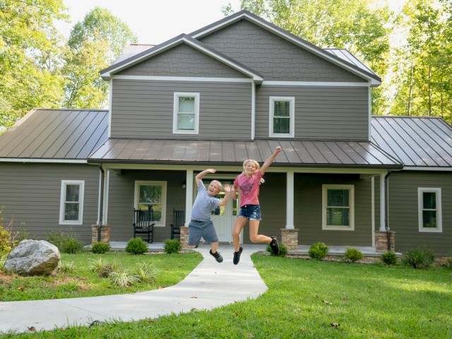 Children are jumping in front of a modern grey house with a lush green yard and trees in the background.