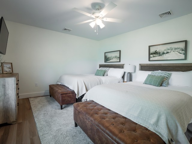 A clean, modern bedroom with two beds, wooden furniture, artwork, a TV, and a ceiling fan in a bright, minimalist space.