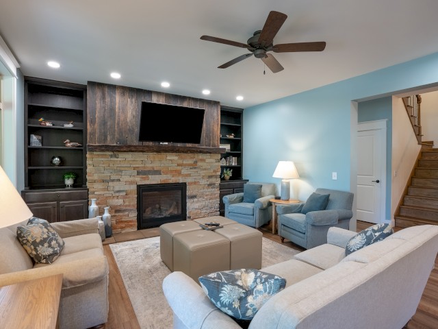 A cozy living room with a fireplace, a mounted TV, several chairs, a beige sofa, and built-in shelves, with hardwood floors and a staircase.