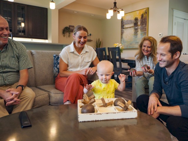 Family enjoys quality time at home with a baby playing with wooden blocks, surrounded by smiling adults in a cozy living room.