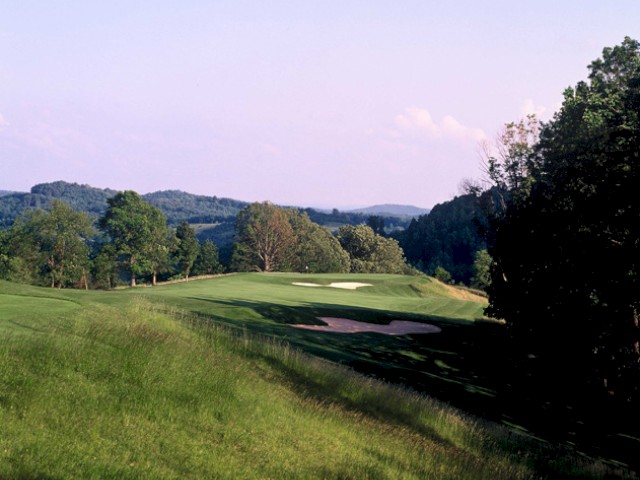 A scenic view of a golf course with green fairways, trees on the right, and distant hills under a clear, light blue sky.