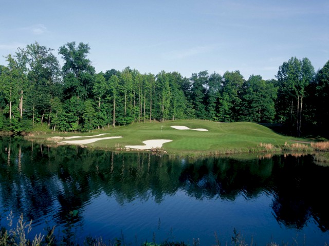 The image shows a lush golf course with a green, sand traps, a large pond in the foreground, and trees in the background beneath a clear blue sky.