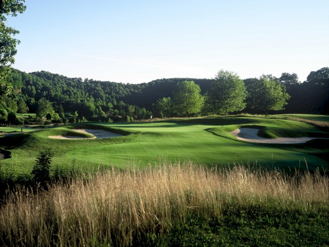 A lush golf course with neatly trimmed grass, sand bunkers, trees, and a backdrop of green hills and a clear blue sky.