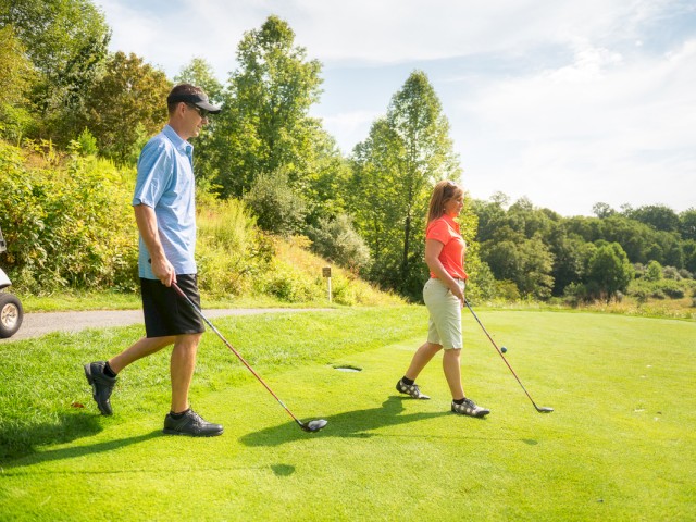 Two people are on a golf course, each holding a golf club, with a golf cart in the background amidst lush greenery.