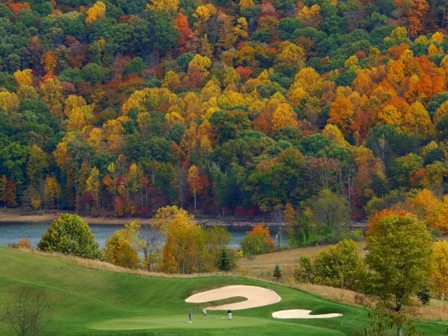 A golf course set against a backdrop of colorful autumn trees, with golfers playing near sand traps and a small lake nearby.