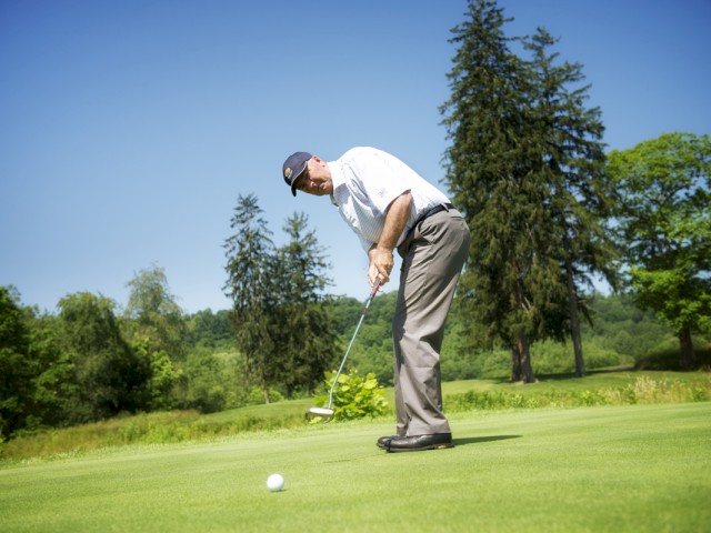 A man is putting on a golf course surrounded by trees and greenery on a clear, sunny day, focusing on his shot.