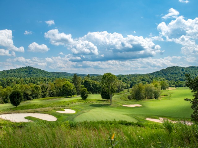 A scenic view of a golf course with green fairways, sand bunkers, lush trees, and rolling hills under a partly cloudy blue sky.