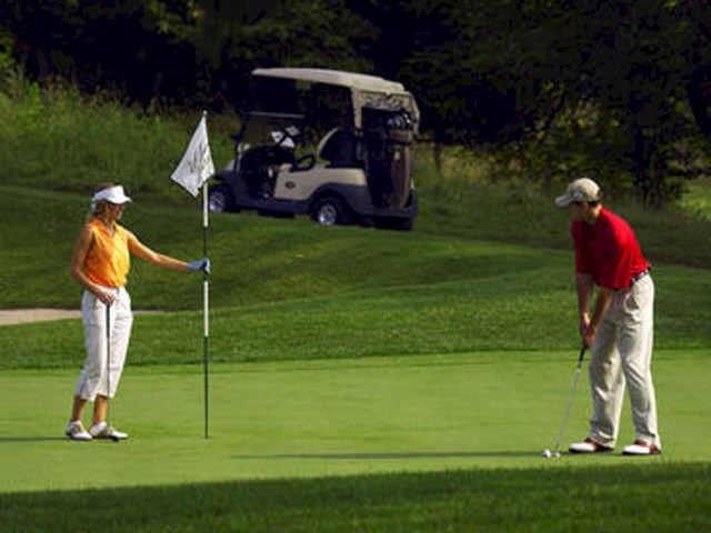 A woman is holding a flag on a golf course while a man is putting. A golf cart is parked in the background.