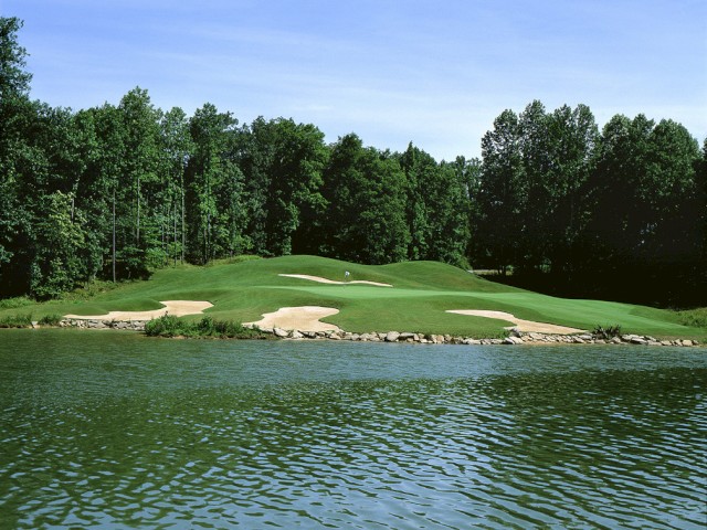 A scenic golf course features a green surrounded by sand traps, bordered by a pond and lush forest under a clear blue sky.