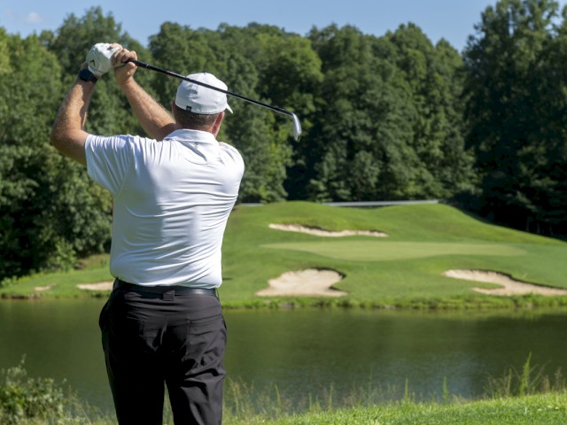 A golfer in a white shirt and hat swings his club on a course, aiming towards the green, with sand traps and trees in the background.